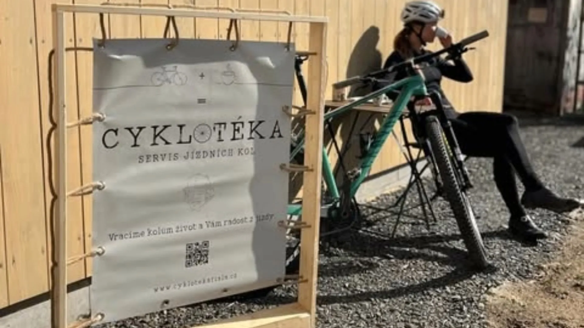 Cyklistka odpočívá u cedule Cyklotéka před dílnou ve Veselí